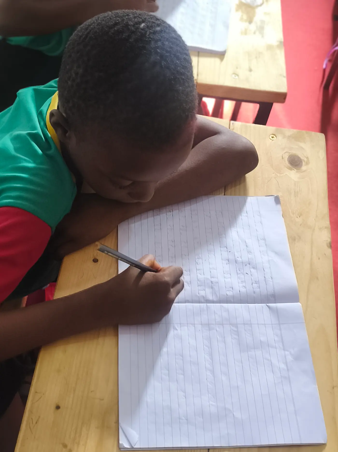 Menino de uniforme verde e vermelho escrevendo em caderno pautado sobre a carteira