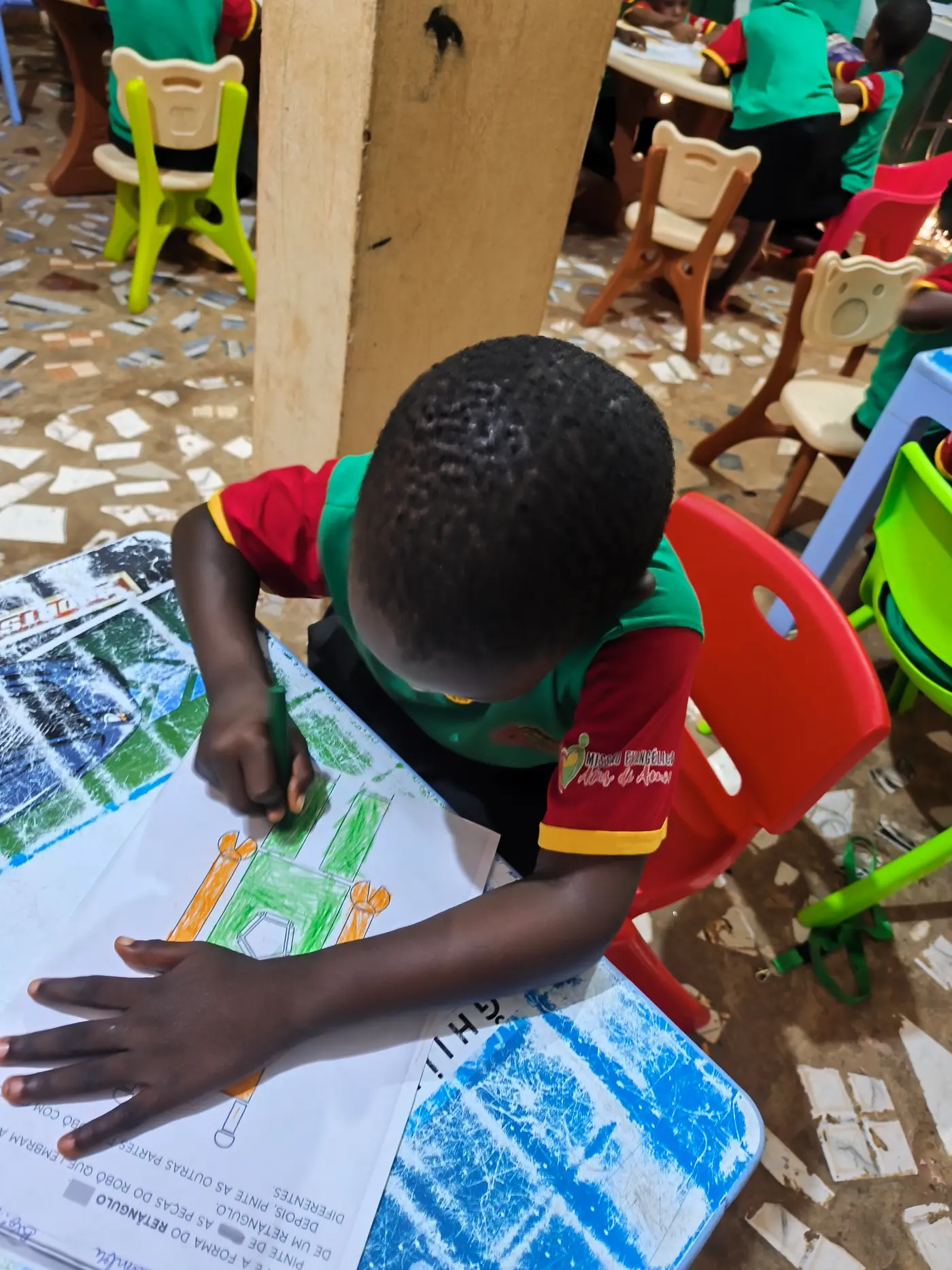 Menino colorindo uma atividade de formas geométricas sobre a mesa, cadeiras coloridas ao redor