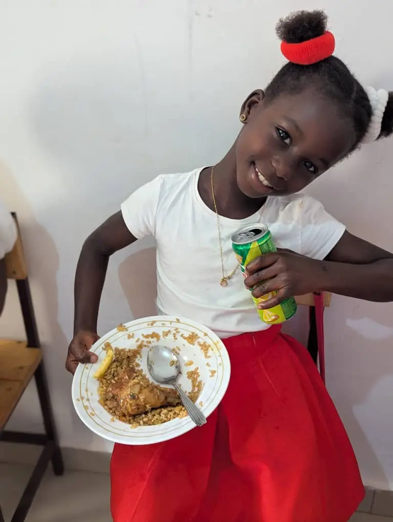 Menina de camisa branca e saia vermelha segurando uma lata de refrigerante e um prato de arroz com frango